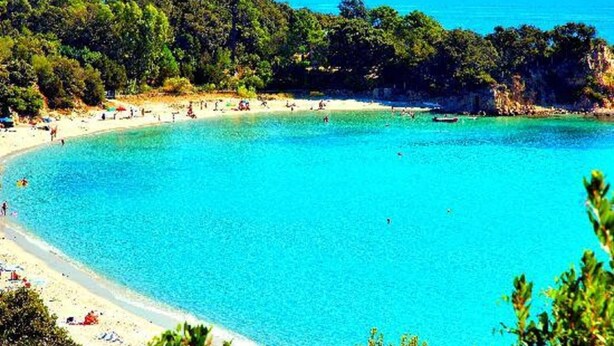 Uma praia com águas turquesa cristalinas, areal arenoso e vegetação verde exuberante ao fundo.