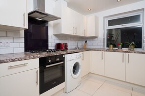 A modern kitchen with white cabinetry, a stainless steel range hood, a red toaster, a Bosch washing machine, and a window with plants. - Spacious 7 Double bedroom House for large groups (London)