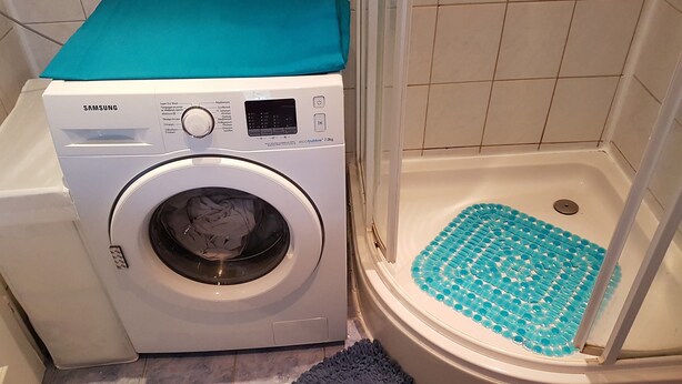 A front-loading Samsung washing machine with a control panel, a blue mat in the shower, and a grey rug on the floor. - Gikas Apartments  - Superior  Suite - 100 meters from the beach  (Kerkyra)