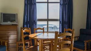 A room with a wooden dining table and chairs, a television on a wooden cabinet, and a large window with curtains. - Kolbast Apartment, balcony, garage (Armação de Pêra)