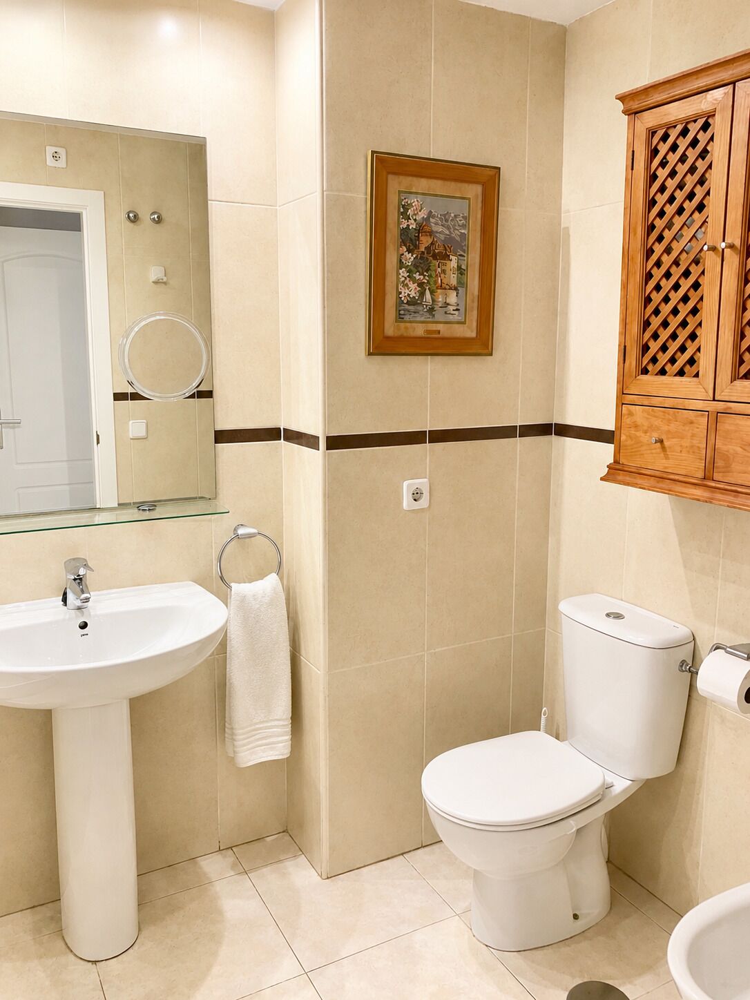 Un bagno con lavandino, WC e specchio. In parete, un quadro incorniciato e un armadietto in legno sopra il water.
