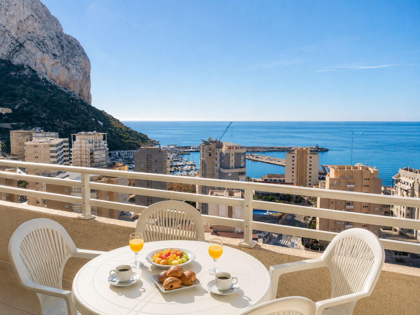 Un balcon avec une table préparée pour le petit-déjeuner, offrant une vue sur le paysage urbain côtier et la mer.