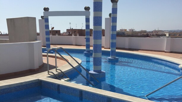A rooftop swimming pool with blue-tiled edges and white pillars, surrounded by a white wall and a clear sky.