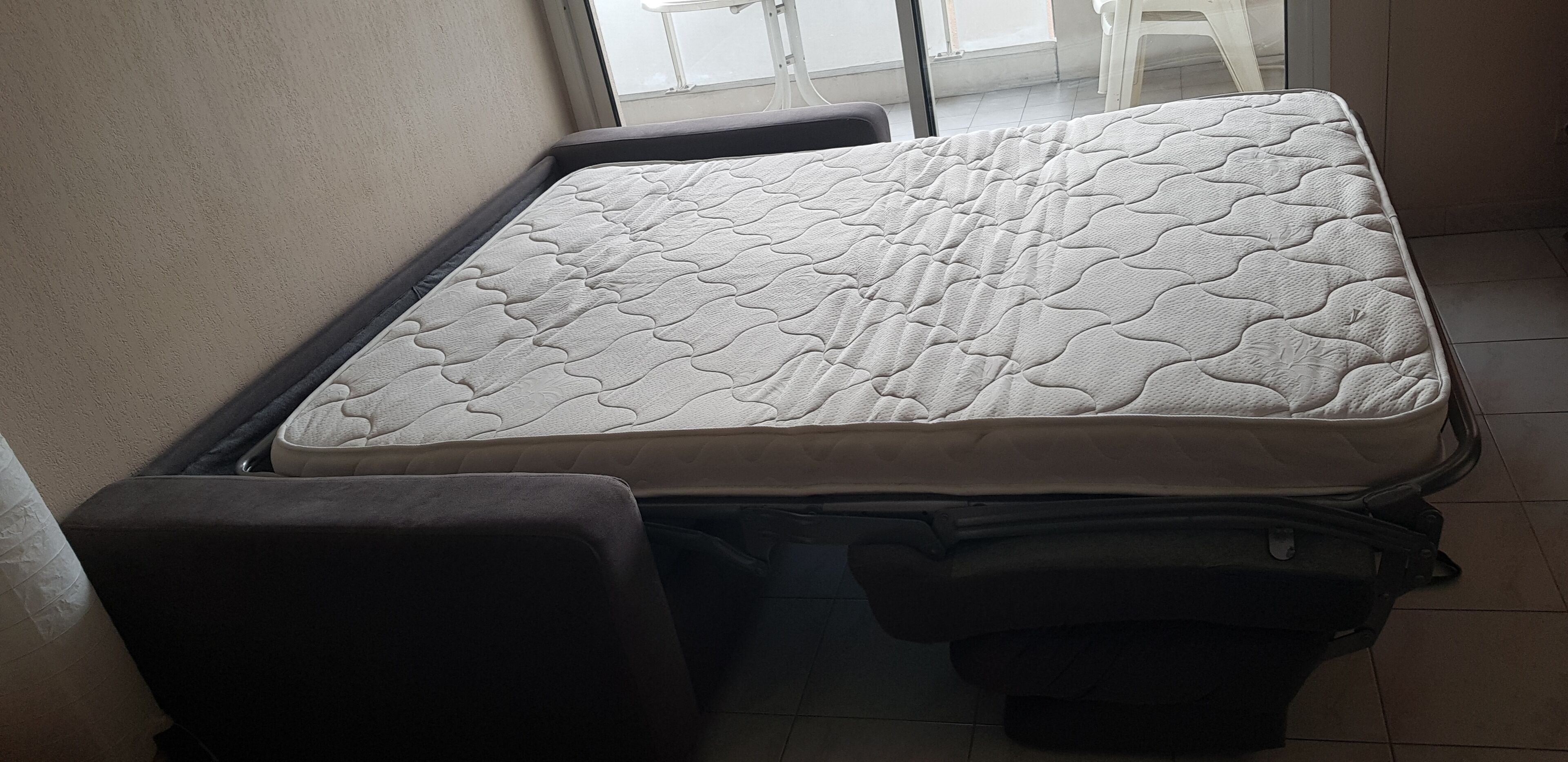 Una cama compacta con colchón acolchado en una habitación pequeña con suelo de baldosas y una ventana.
