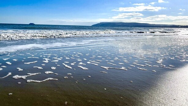 A beach with clear water, a distant ship, and a hilly landscape.