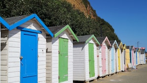A row of colorful beach huts with doors, some with numbers above them, along a sandy beach with a cliff in the background.