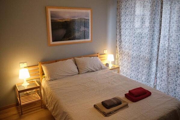 A neatly made bed with a patterned bedspread, two bedside lamps, a framed picture on the wall, and a window with patterned curtains.