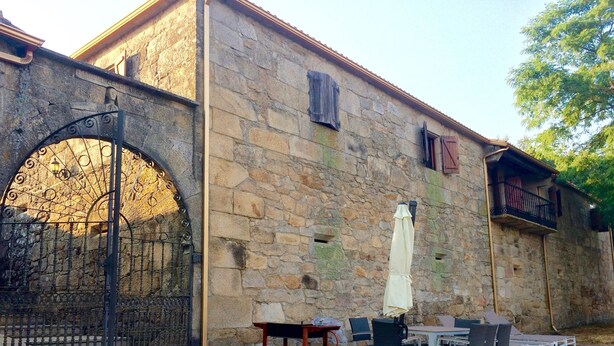 A stone building with a large arched metal gate, outdoor seating with tables and chairs, and a small table with a chair. - Bed Century XII - Pontevedra (Ribeira Sacra) (Fornas - Pontevedra)