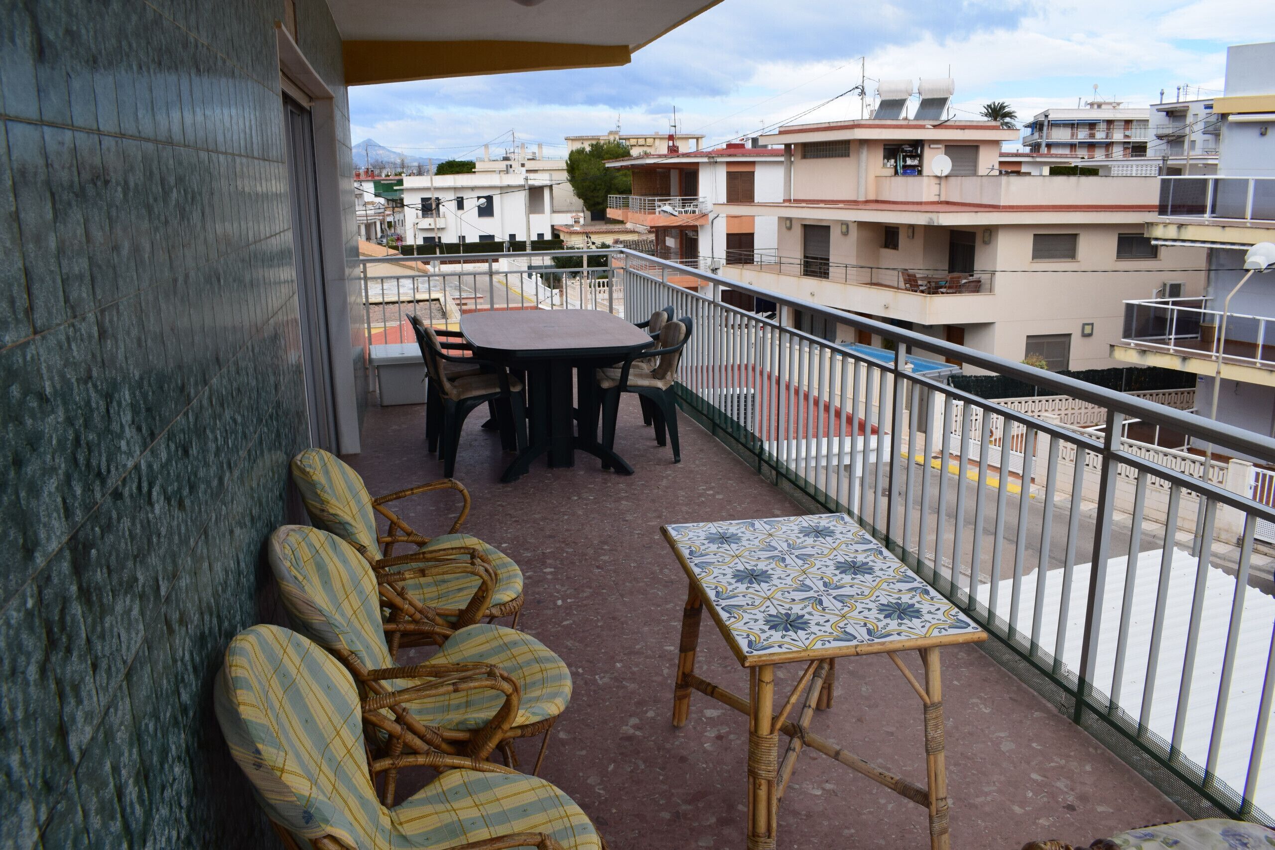 A balcony with wicker chairs and a table, overlooking a residential area with multiple buildings.