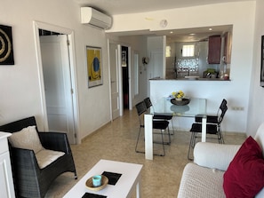 A modern living room with a glass coffee table, a sofa, and a dining area with a glass table and chairs. There is a kitchen visible in the background with a window and cabinets.