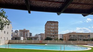 A swimming pool with a surrounding grassy area, a pergola overhead, and a backdrop of residential buildings. - Beautiful apartment on the beachfront (PLAYA-GRAU DE GANDÍA)