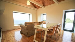 Quarto com mesa de jantar de vidro, piso de madeira, sofá bege e grandes janelas com vista para o mar.