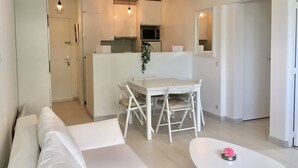 Un salon moderne et minimaliste, avec un canapé blanc, une table à manger et des chaises, un coin cuisine équipé d’un four à micro-ondes et un luminaire suspendu.