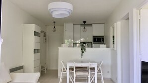 Un salon moderne et minimaliste, avec un canapé blanc, une table à manger et des chaises, ainsi qu’un coin cuisine doté de meubles et d’appareils électroménagers blancs.
