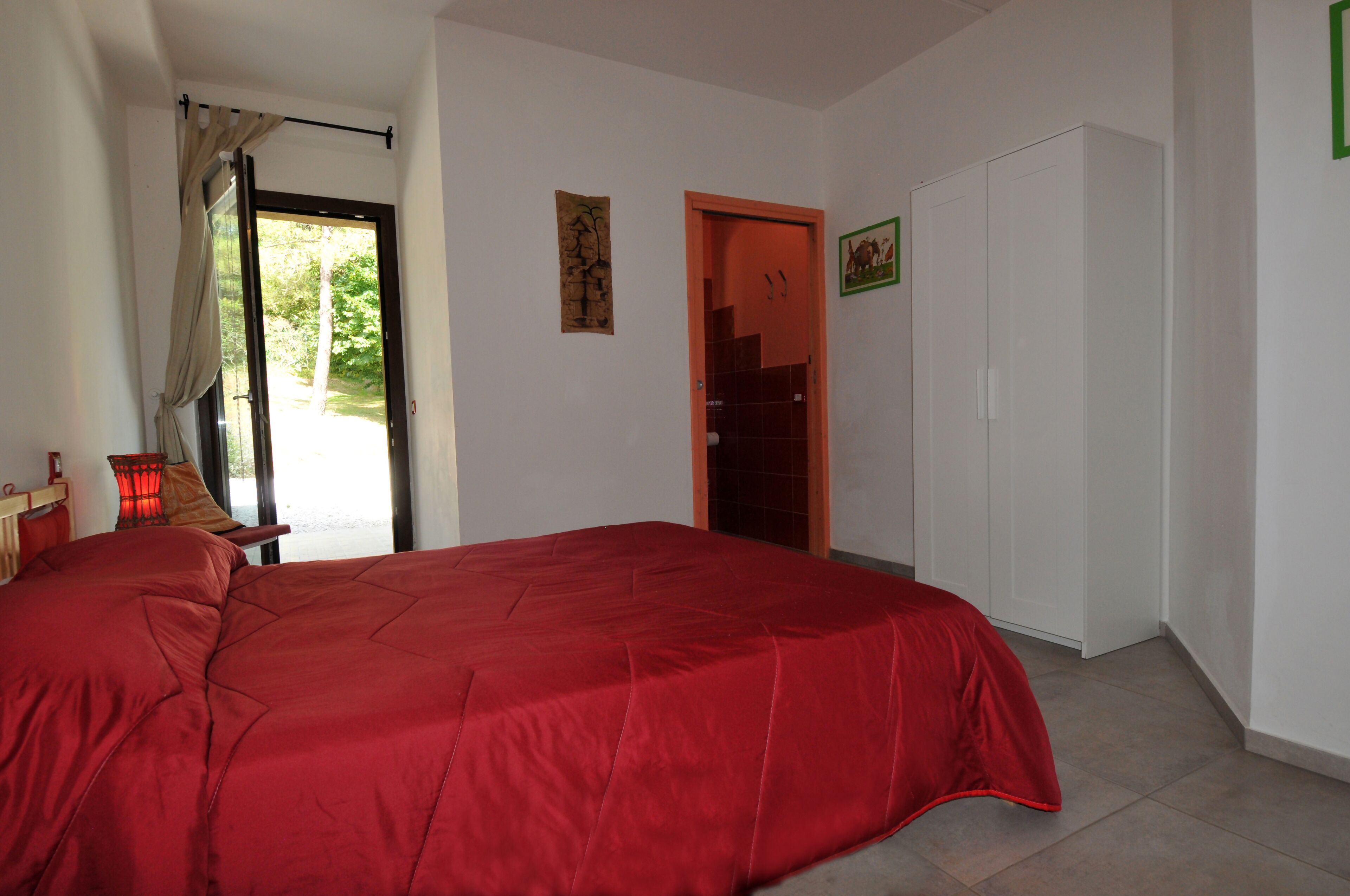 A bedroom with a large bed, a wardrobe, a door leading outside, and a small table with a lamp.