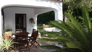 Une terrasse couverte, agrémentée d’une table et de chaises, entourée d’une végétation luxuriante et bordée d’un bâtiment blanc au toit recouvert de tuiles.