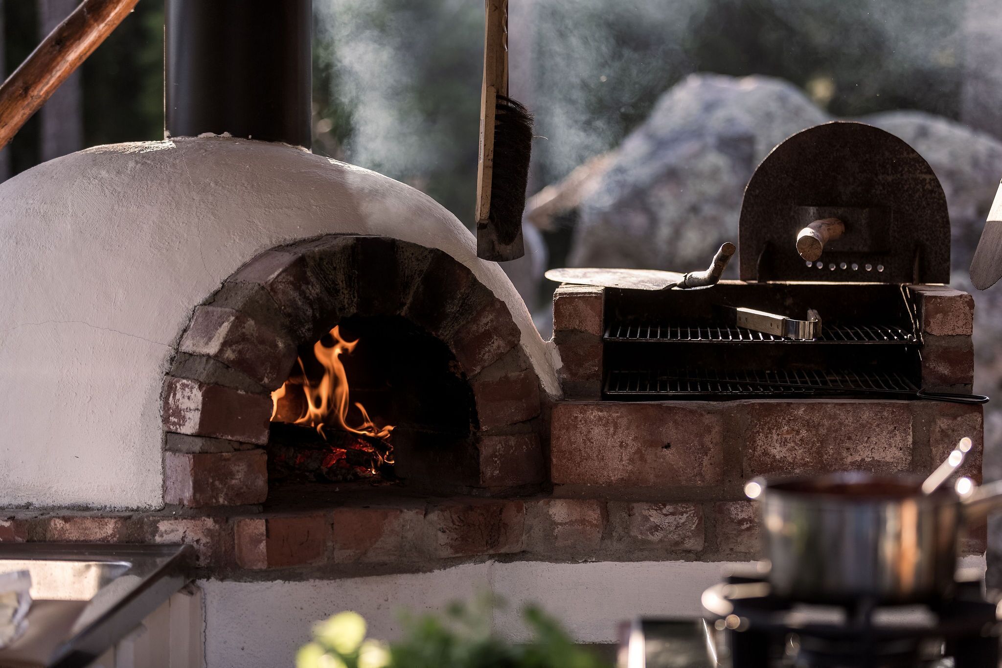 A brick oven pizza oven with a fire burning inside, a grill on top, and a pot on a stove nearby.