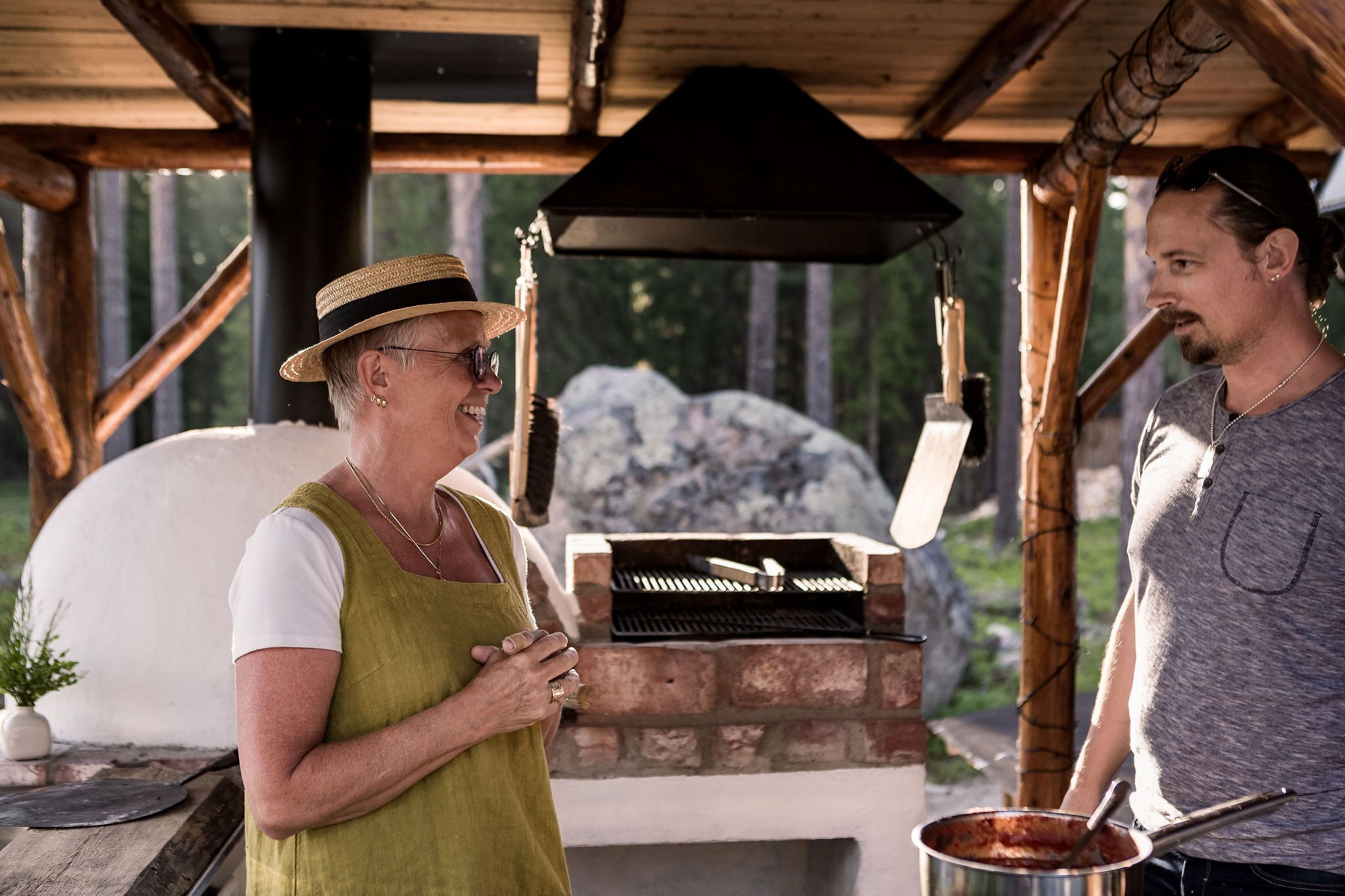 A person in a green apron and straw hat standing next to a brick oven, with another person in a grey shirt nearby.