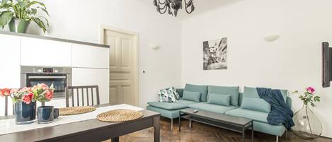 En moderne stue med sofa, sofabord og spisebord. Der er et køkkenområde med ovn og en potteplante.