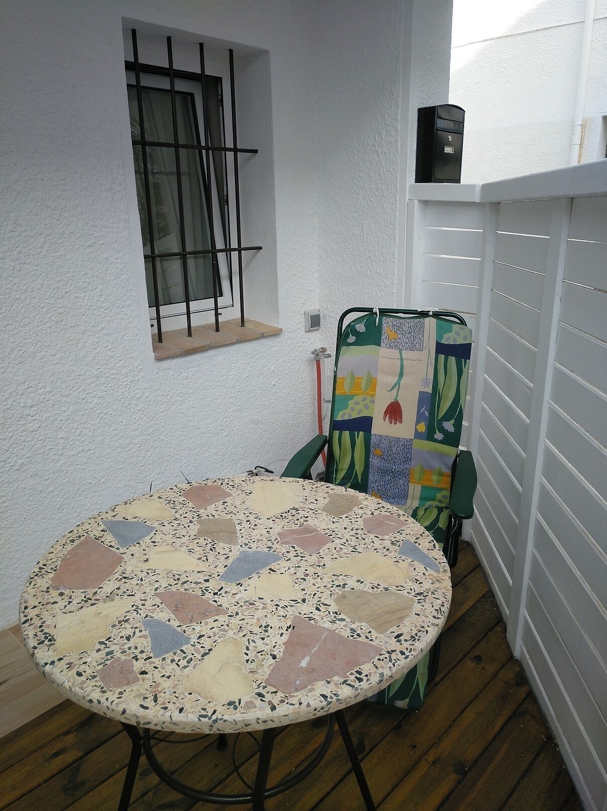 A small room with a round table, a window with bars, a chair with a colorful cushion, and a small shelf with a box.
