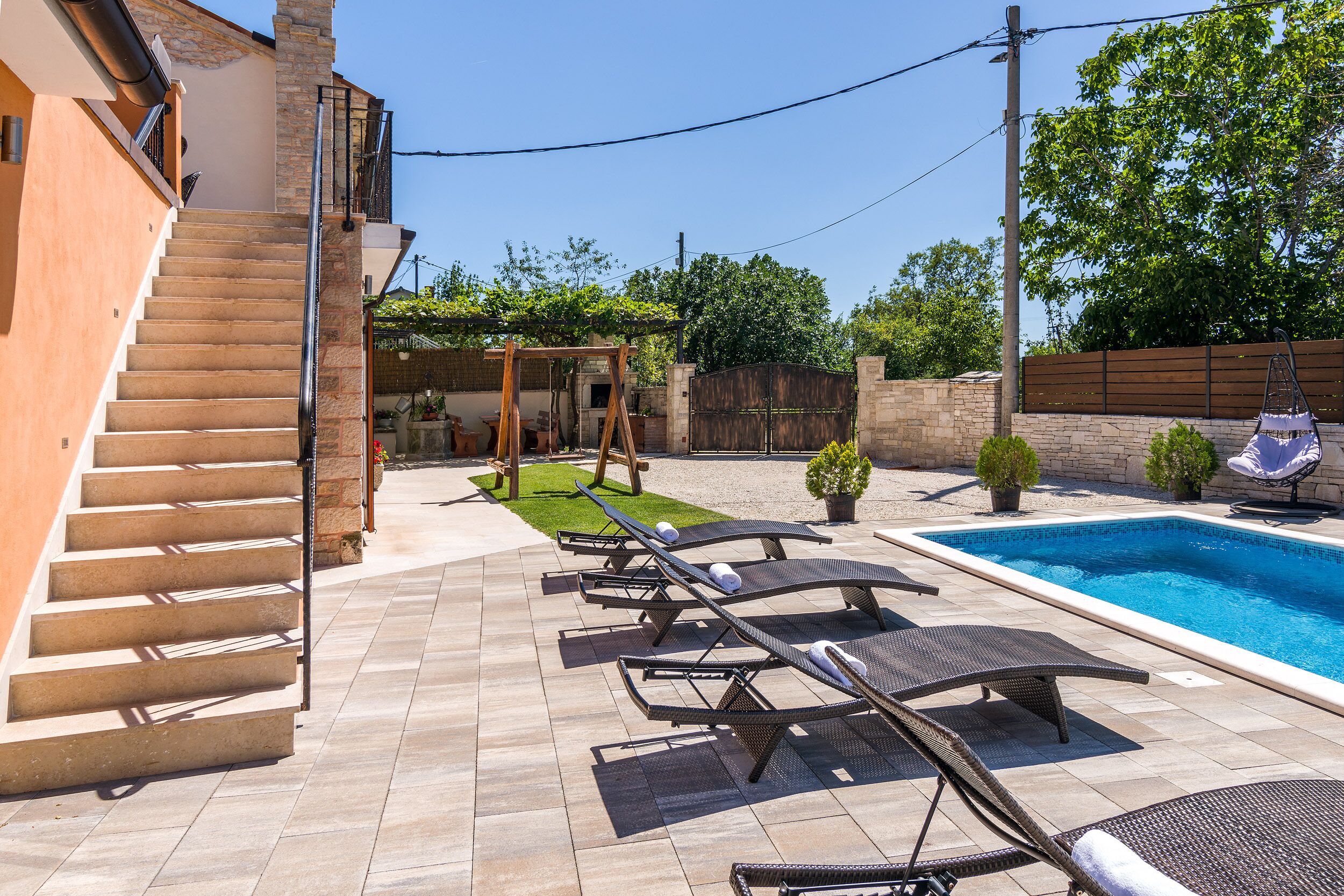 A backyard with a swimming pool, lounge chairs, a wooden swing set, and a staircase leading to a second floor.