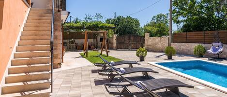 A backyard with a swimming pool, lounge chairs, a wooden swing set, and a staircase leading to a second floor.