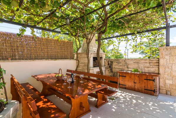 Um pátio externo com mesa e bancos de madeira, uma parede de pedra e um forno de tijolos.