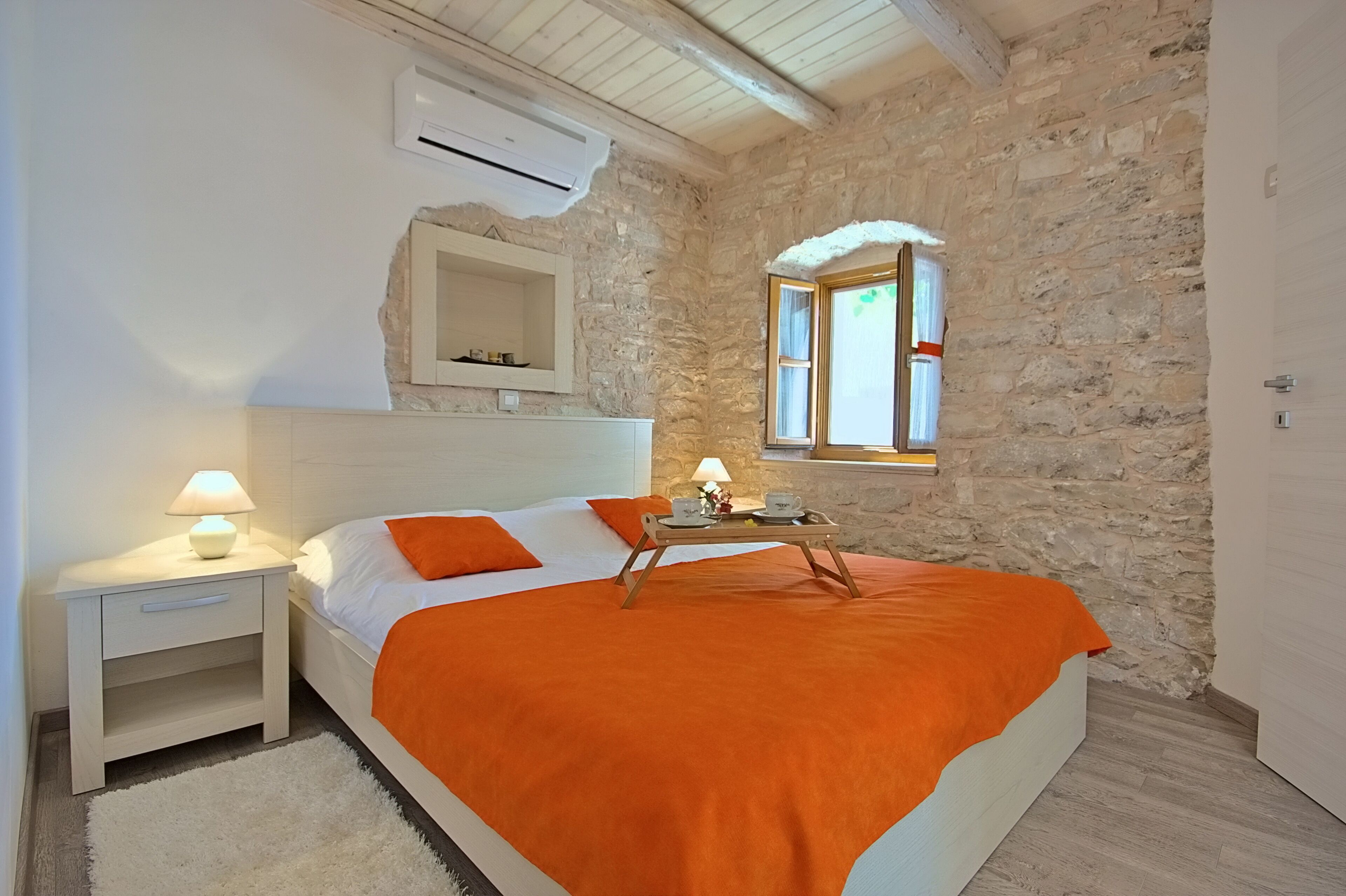 Ein Schlafzimmer mit Steinwand, einem Bett mit orangefarbener Decke, einem Nachttisch mit Lampe und einem Fenster mit Vorhängen.