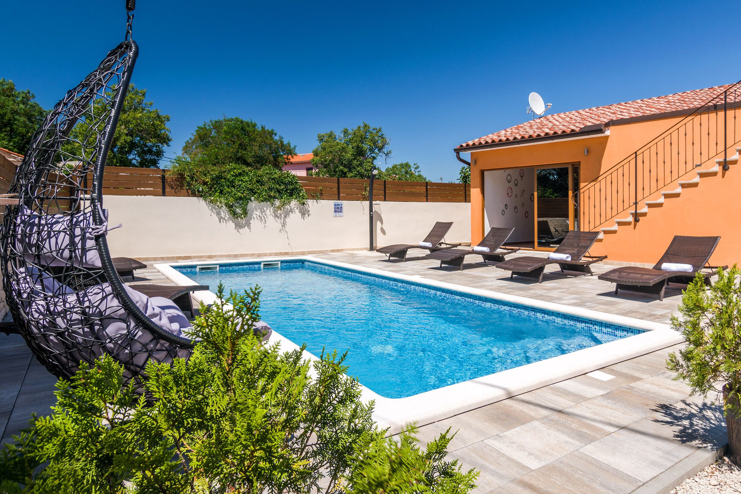 A swimming pool with lounge chairs, a hanging chair, and a small building with a staircase.
