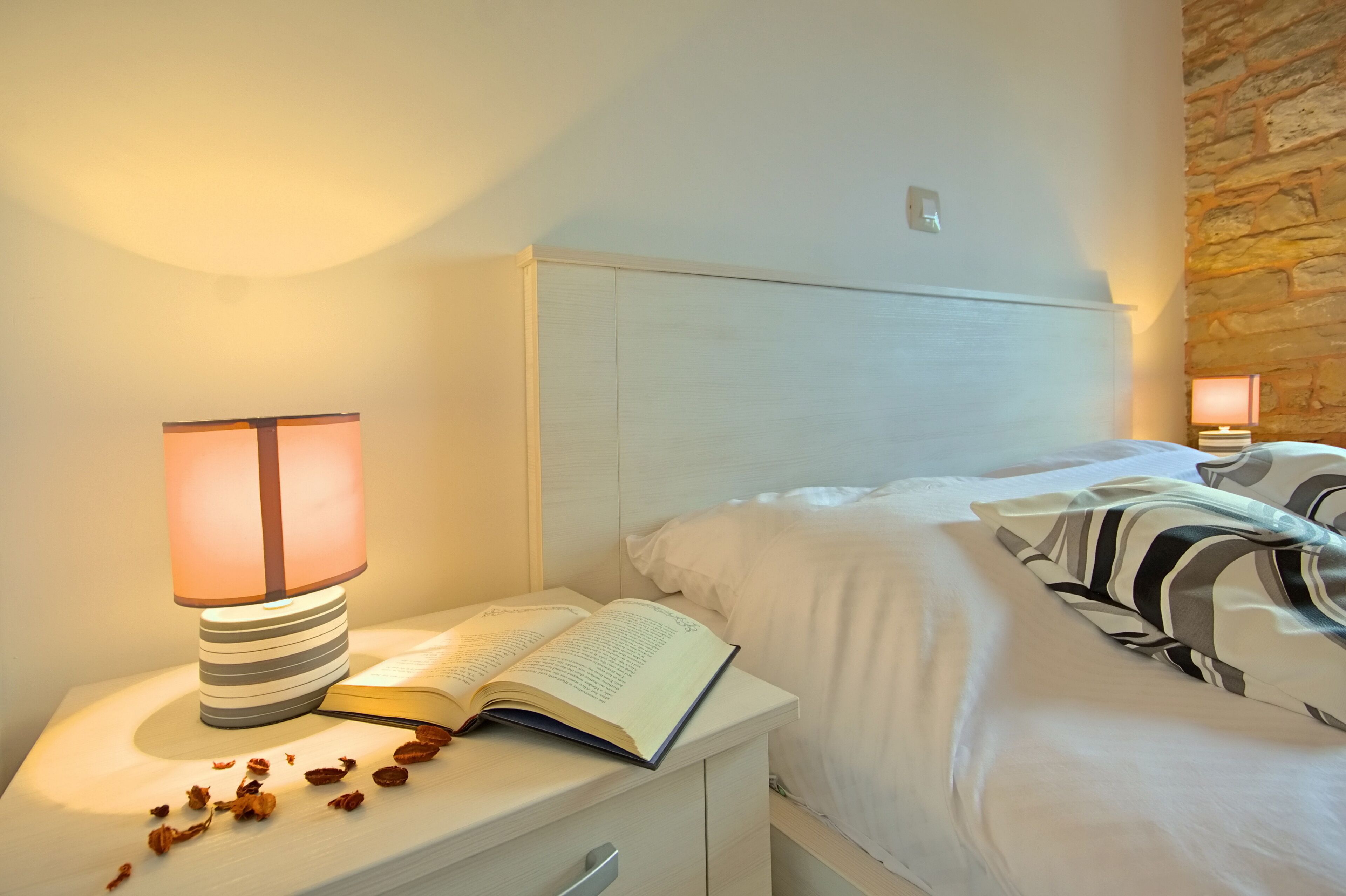 A neatly made bed with white bedding and striped pillows, a bedside table with a lamp, and an open book.