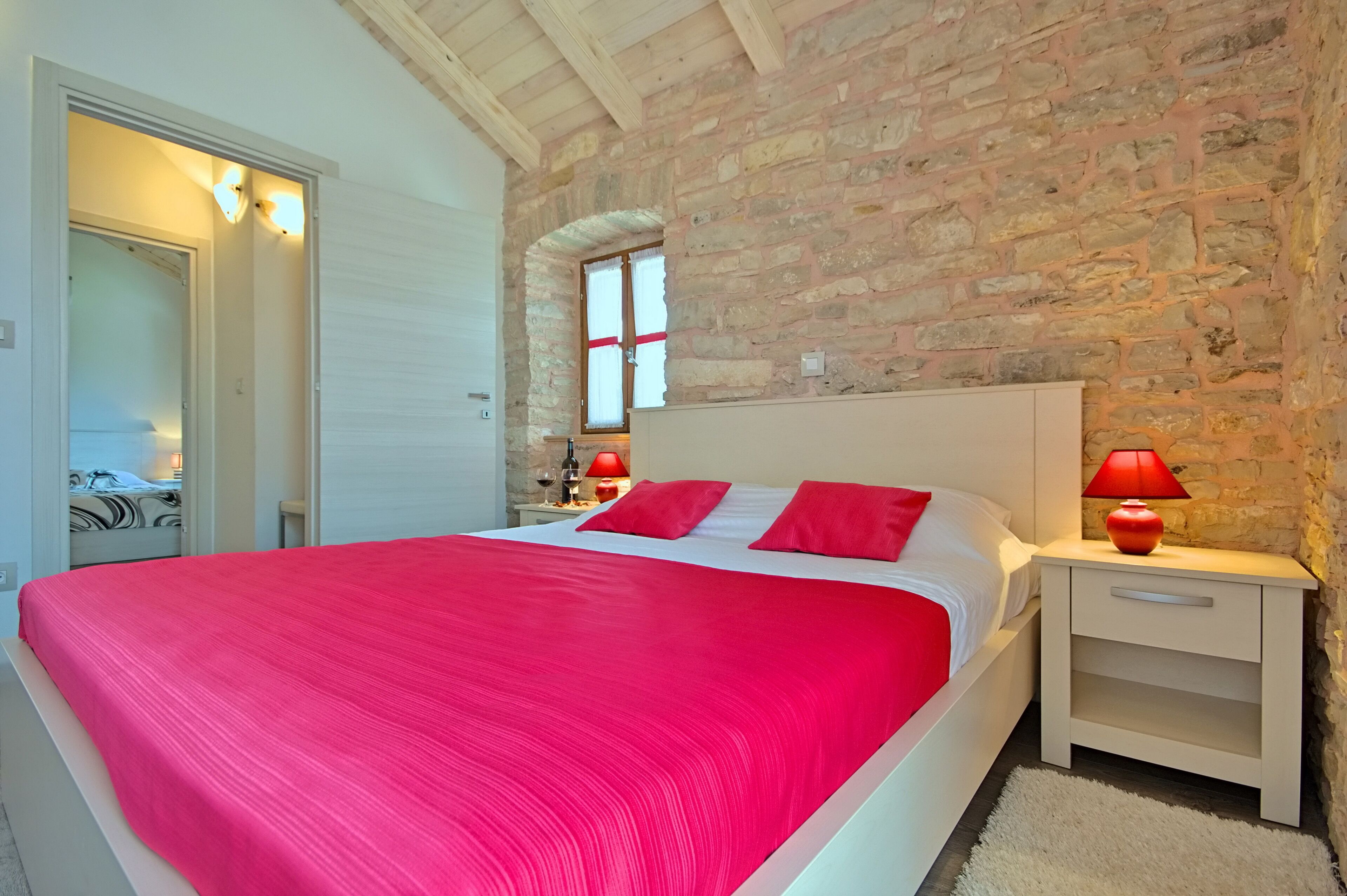 Un dormitorio con pared de piedra, cama con ropa de cama roja, mesita de noche con lámpara roja y ventana con cortinas.