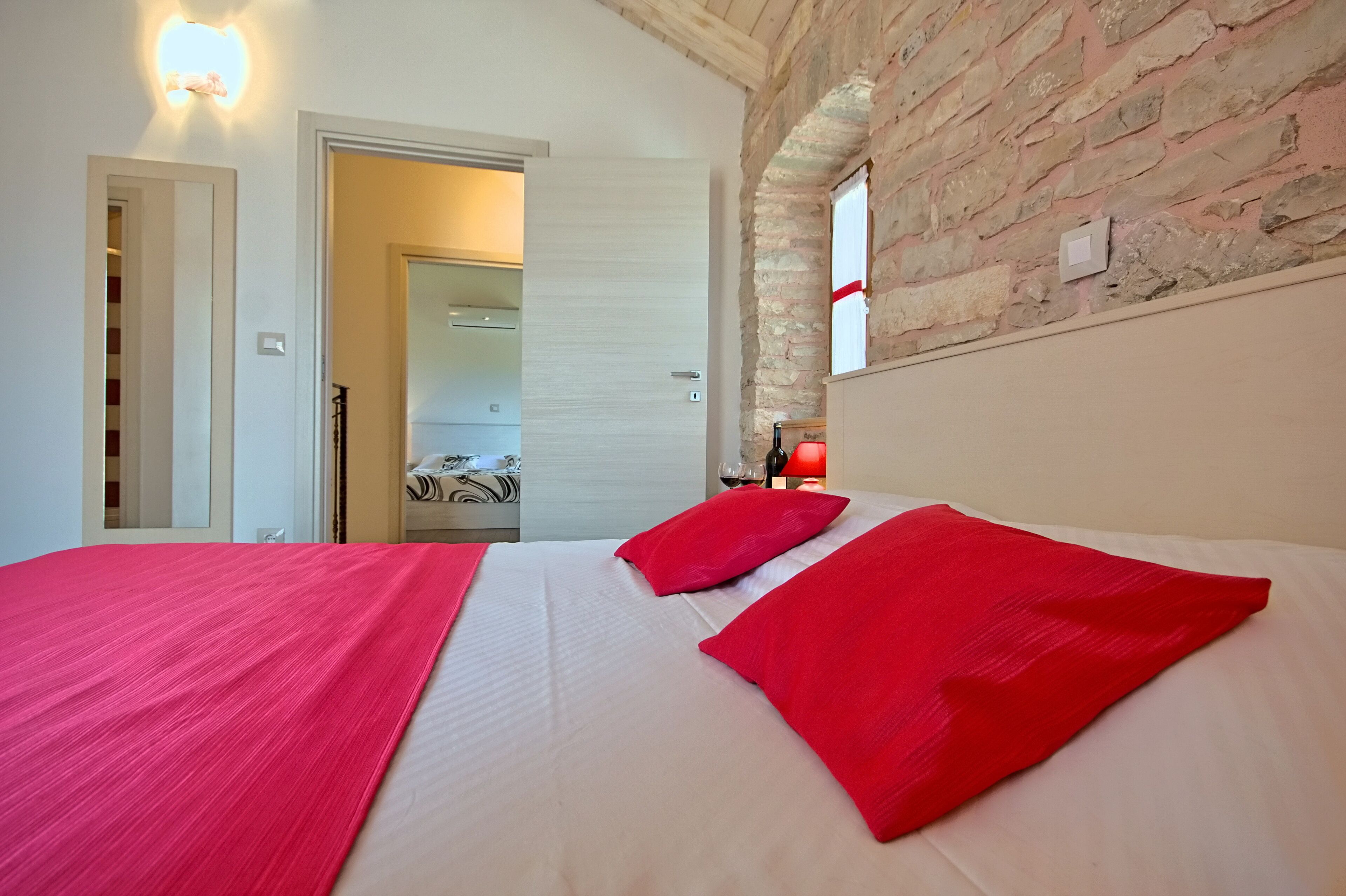 A bedroom with a large bed, red pillows, a brick wall, and a door leading to another room.