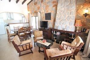 Ein gemütliches Wohnzimmer mit Steinwand, Kamin und Fernseher. Holzstühle und ein Tisch mit Unterlage sind ebenfalls vorhanden.