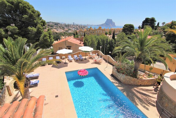 Ein Swimmingpool mit Liegestühlen und Sonnenschirmen, umgeben von Palmen und Blick auf Stadt und Meer.