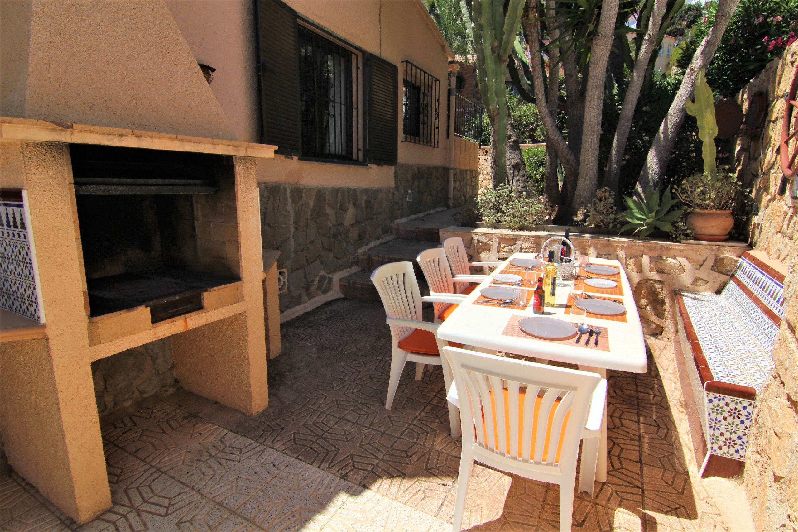 Une terrasse extérieure avec un sol carrelé en pierre, un barbecue, des chaises blanches et une table à manger pour quatre personnes.