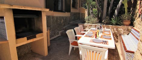 An outdoor patio with a stone-tiled floor, a grill, white chairs, and a dining table set for four.
