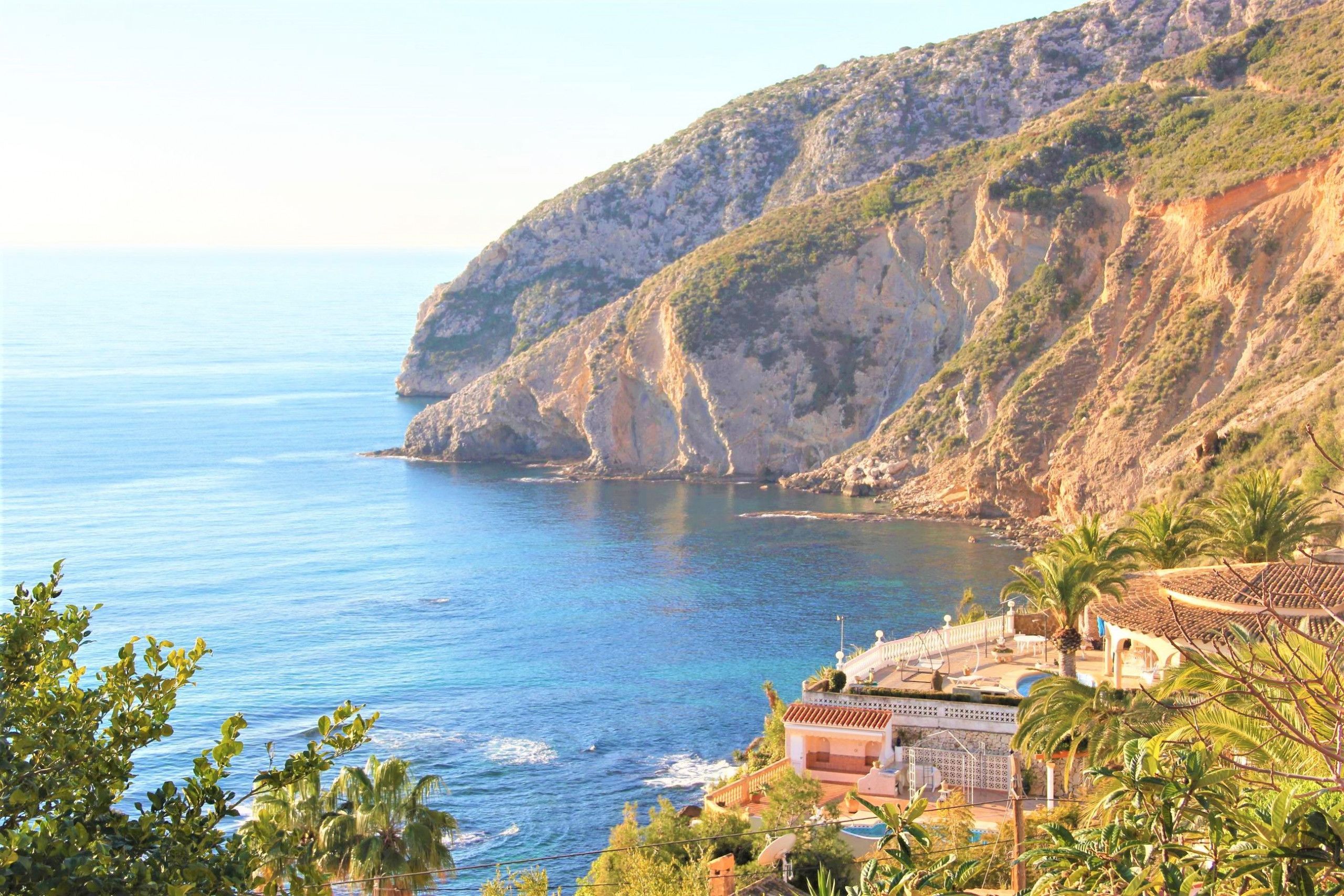 Une propriété située sur la côte, offrant une vue sur la mer, avec des montagnes en arrière-plan et un bâtiment doté d’un toit en tuiles.