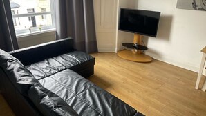 A modern living room with a black leather sofa, a flat-screen TV on a wooden stand, and a window with curtains. - Elderpark Apartment 4 (Glasgow)