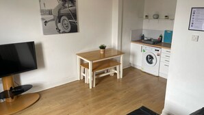 A living room with a TV, a wooden table, and a bench. There is a kitchen area with a washing machine and a sink. - Elderpark Apartment 4 (Glasgow)
