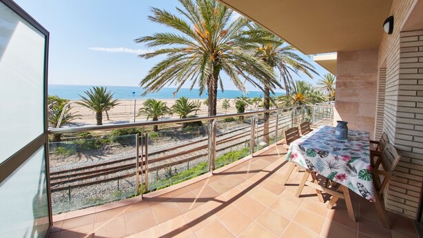 Un balcone con tavolo e sedie, affacciato su una spiaggia con palme e cielo sereno.