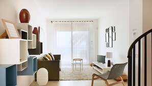 Un soggiorno moderno con un divano, un tavolino e una televisione. Si vedono scaffali con oggetti decorativi e una scala sullo sfondo.