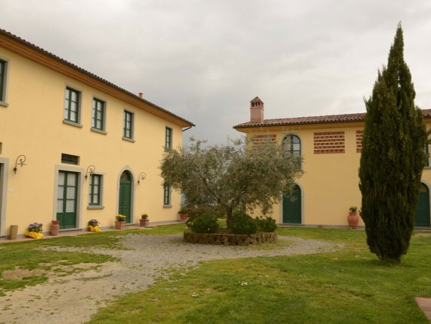 Ein Innenhof mit einem kleinen Baum, einer hohen Zypresse und einem Gebäude mit gelber Fassade und grünen Türen.