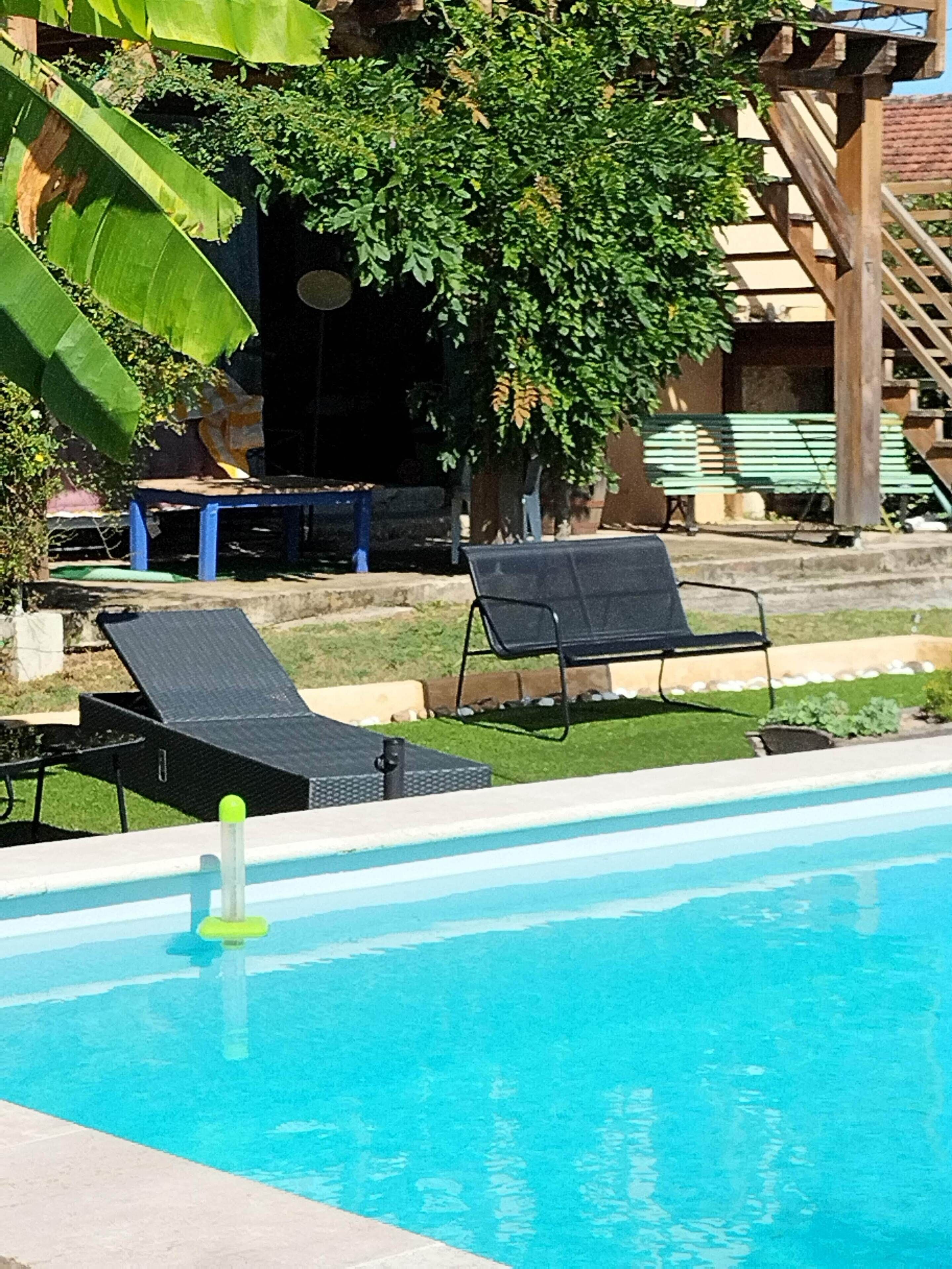 Una piscina con sillas de descanso, una pérgola de madera y un árbol al fondo.