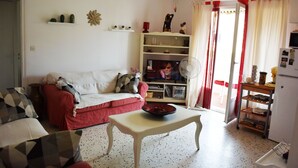 A living room with a sofa, a coffee table, and a television. There is a fan, a shelf with decorative items, and a door leading to another room.
