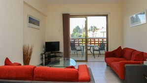 Ein Wohnzimmer mit einem roten Sofa, einem Glastisch, einem Flachbildfernseher und einer Glasschiebetür, die zu einer Außenpatio mit Stühlen und einem Tisch führt.