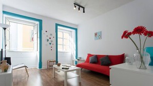 Een moderne woonkamer met een rode bank, een witte salontafel en een klein bijzettafeltje. Er zijn twee ramen met blauwe omlijsting, een muur met ronde decoraties, en een vaas met rode bloemen op een witte console tafel.