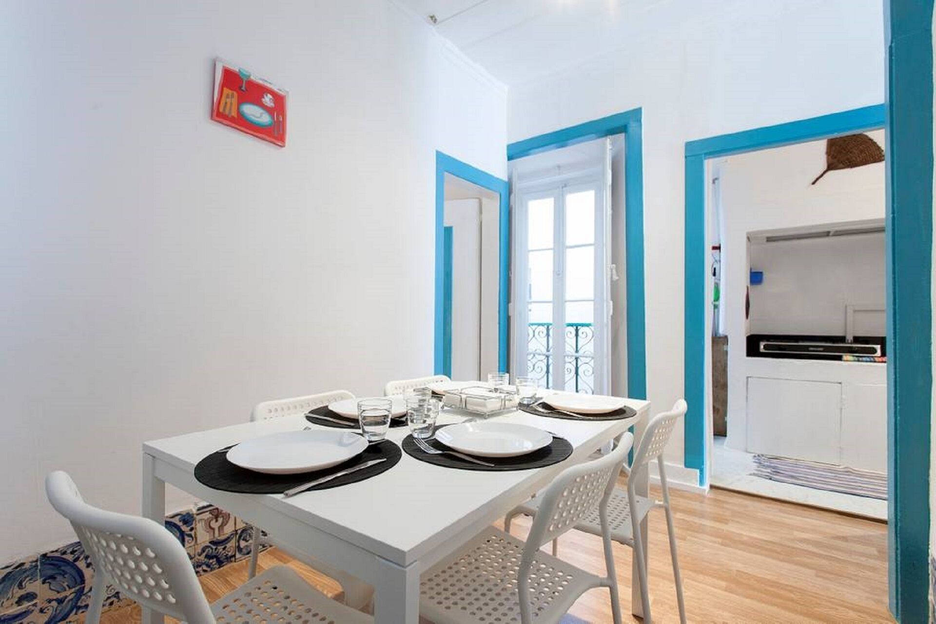 A dining area with a white table set for four, white chairs, and a kitchen visible through an open door.
