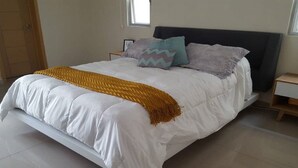 Ein ordentlich gemachtes Bett mit weißer Decke, einer senffarbenen Tagesdecke und zwei Kissen mit geometrischen Mustern.