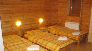 A cabin with three wooden beds, each with a plaid bedspread and a rolled towel. There are two wall-mounted lights and a window with shutters.