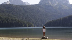 A person standing on a rock by a calm lake with mountains in the background.